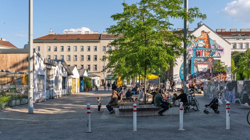 copyright: Moritz Nachtschatt Menschen sitzen auf Bänken rund um Bäume auf einem großteils asphaltierten Platz, daneben und dahinter befinden sich kleinere und größere Gebäude