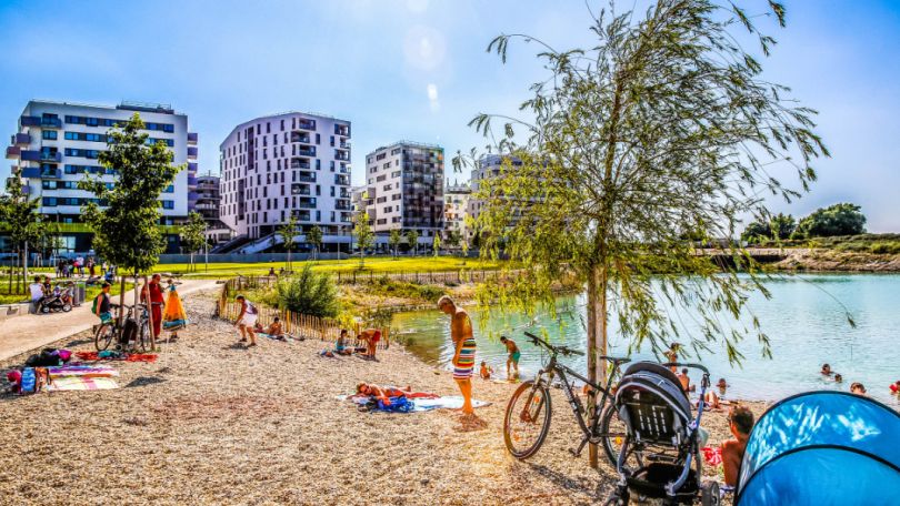 copyright: PID/C. Fürthner Kiesstrand am See, badende Menschen, Gebäude im Hintergrund