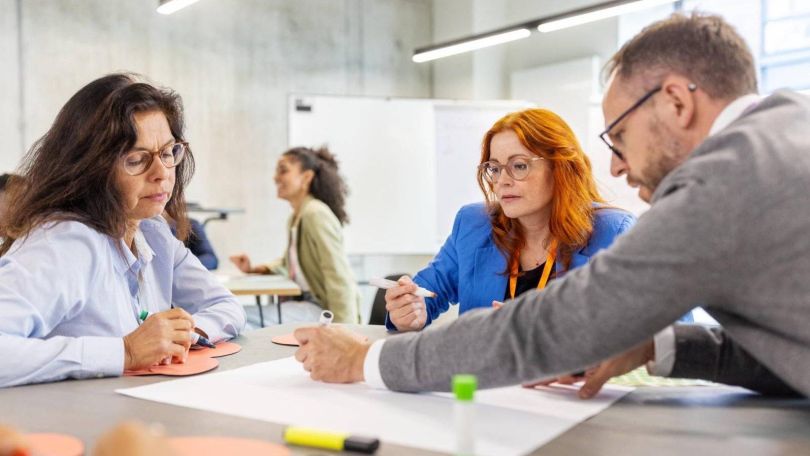 copyright: Getty Images Ein Mann und zwei Frauen sitzen an einem Tisch und schreiben etwas auf ein Plakat in der Mitte des Tisches. Alle tragen Businesskleidung.