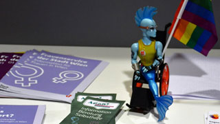 Broschren zu Frauenthemen liegen auf einem Tisch, dazwischen eine Deko-Puppe. Die Puppe hat einen blauen Irkokesen-Haarschnitt und blauen Meerjungfrauschwanz. Sie sitzt in einem Rollstuhl, an dem eine Regenbogenfahne geheftet ist