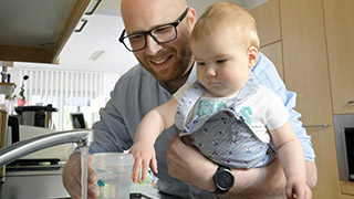Mann mit Baby auf dem Arm fllt eine Trinkflasche am Wasserhahn