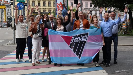 Bezirksvorsteherin Silvia Jankovic und Vertreter*innen der Bezirksvertretung, der HOSI und anderer Organisationen halten eine bunte Transpride-Flagge