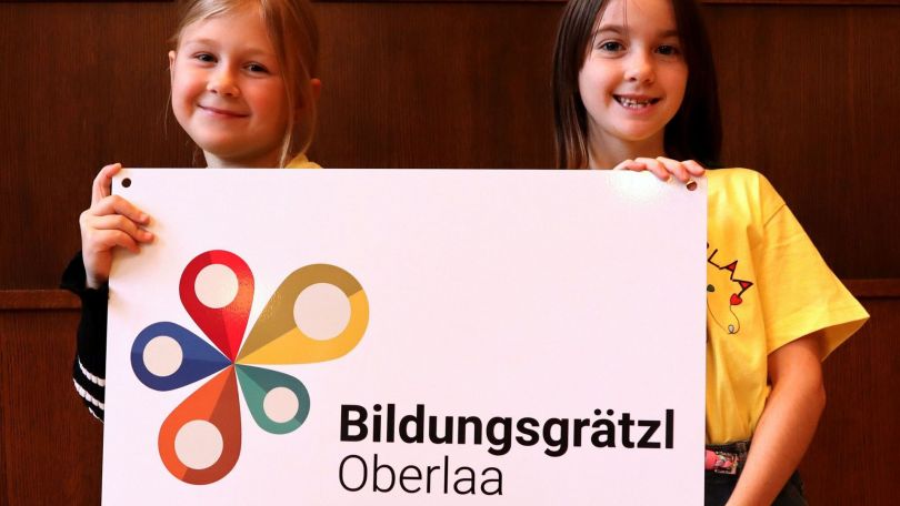 2 Mädchen halten ein Schild mit der Aufschrift "Bildungsgrätzl Oberlaa"