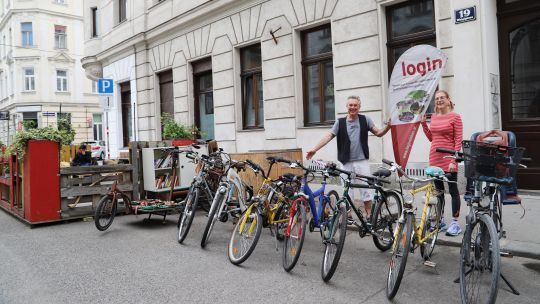 8 bunte Leihfahrräder stehen auf der Straße vor dem Eingang zu "login"