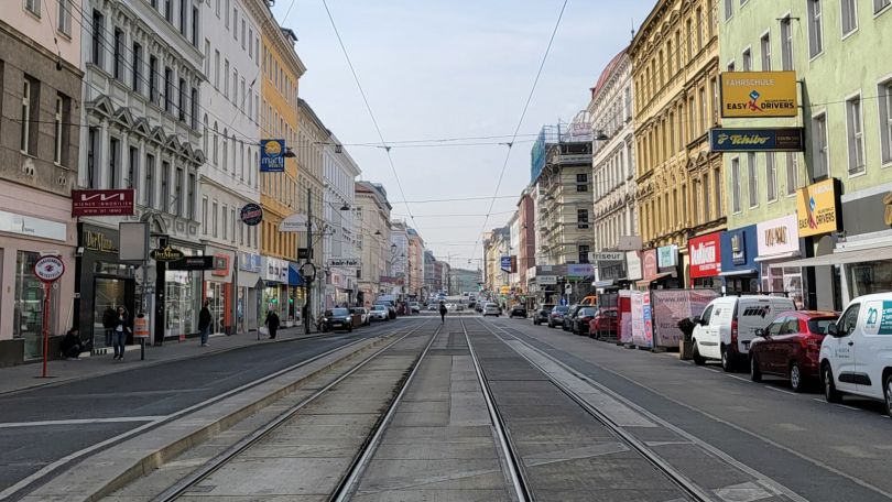 Blick in eine Straße mit Fahrbahn, Schienen, an der Seite parkende Autos und Hausfassaden