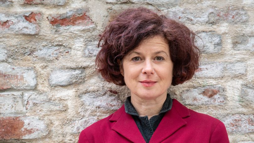 copyright: Kurt Hörbst Frau mit lockigen braunen Haaren und rotem Blazer steht vor einer Backsteinmauer mit verschränkten Händen.