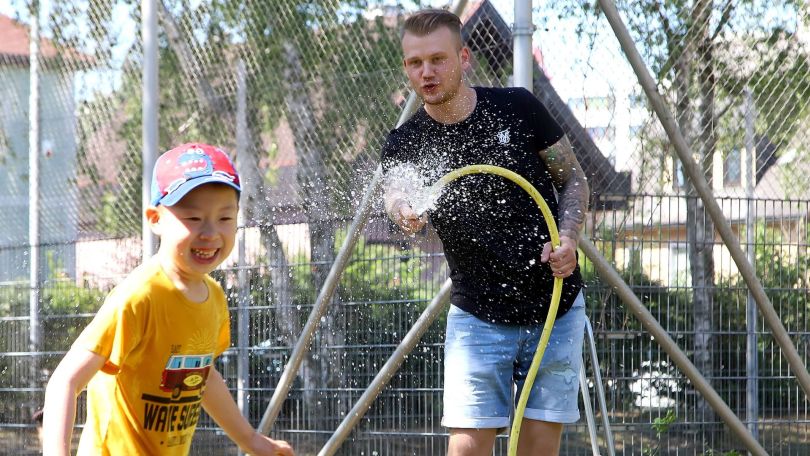 copyright: MA 10 - Wiener Kindergärten/VOTAVA Junger Mann mit schwarzem T-Shirt hält einen Gartenschlauch, vor ihm ein lachendes, laufendes Kind mit gelbem T-Shirt und Kappe.