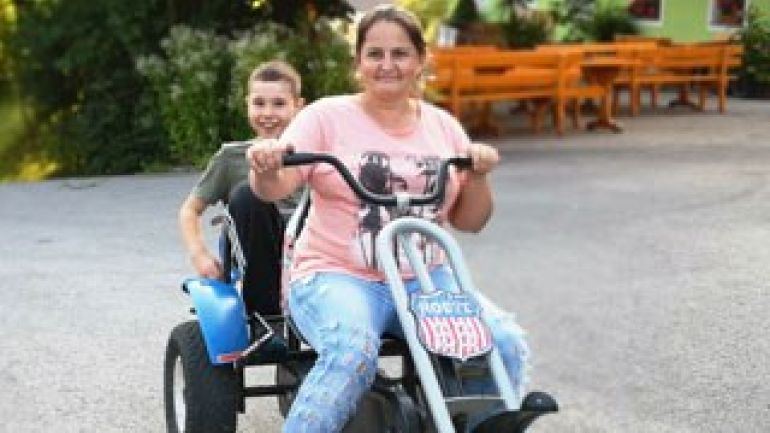 copyright: Wiener Jugenderholung (WIJUG) Eine Mutter sitzt vorne am Lenker eines Gokarts, dahinter sitzt ein lachender Bub.