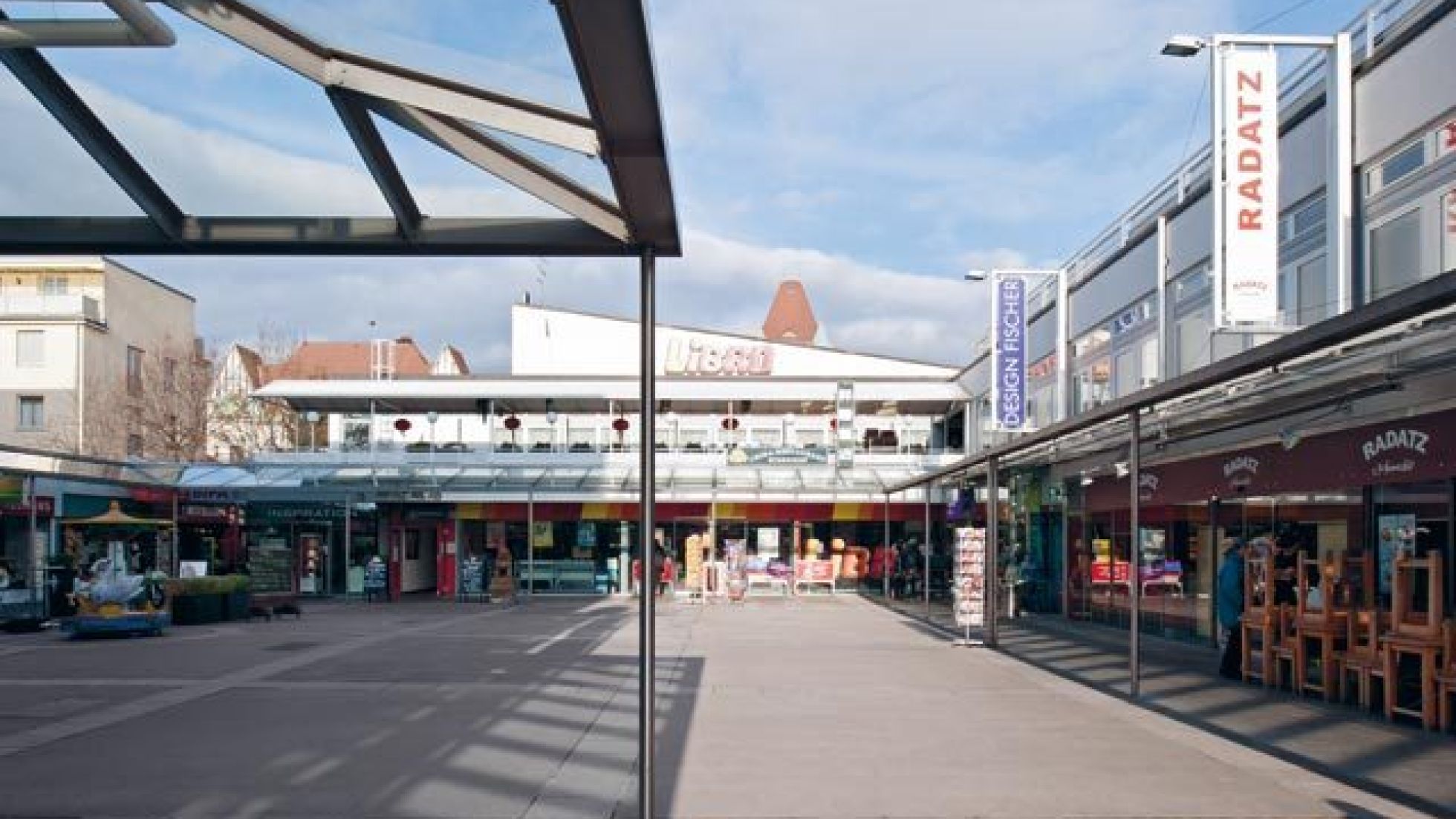 copyright: ©Pez Hejduk befestigter Freibereich, umgeben von Gebäuden mit Geschäften im Erdgeschoß und Glasvordächern