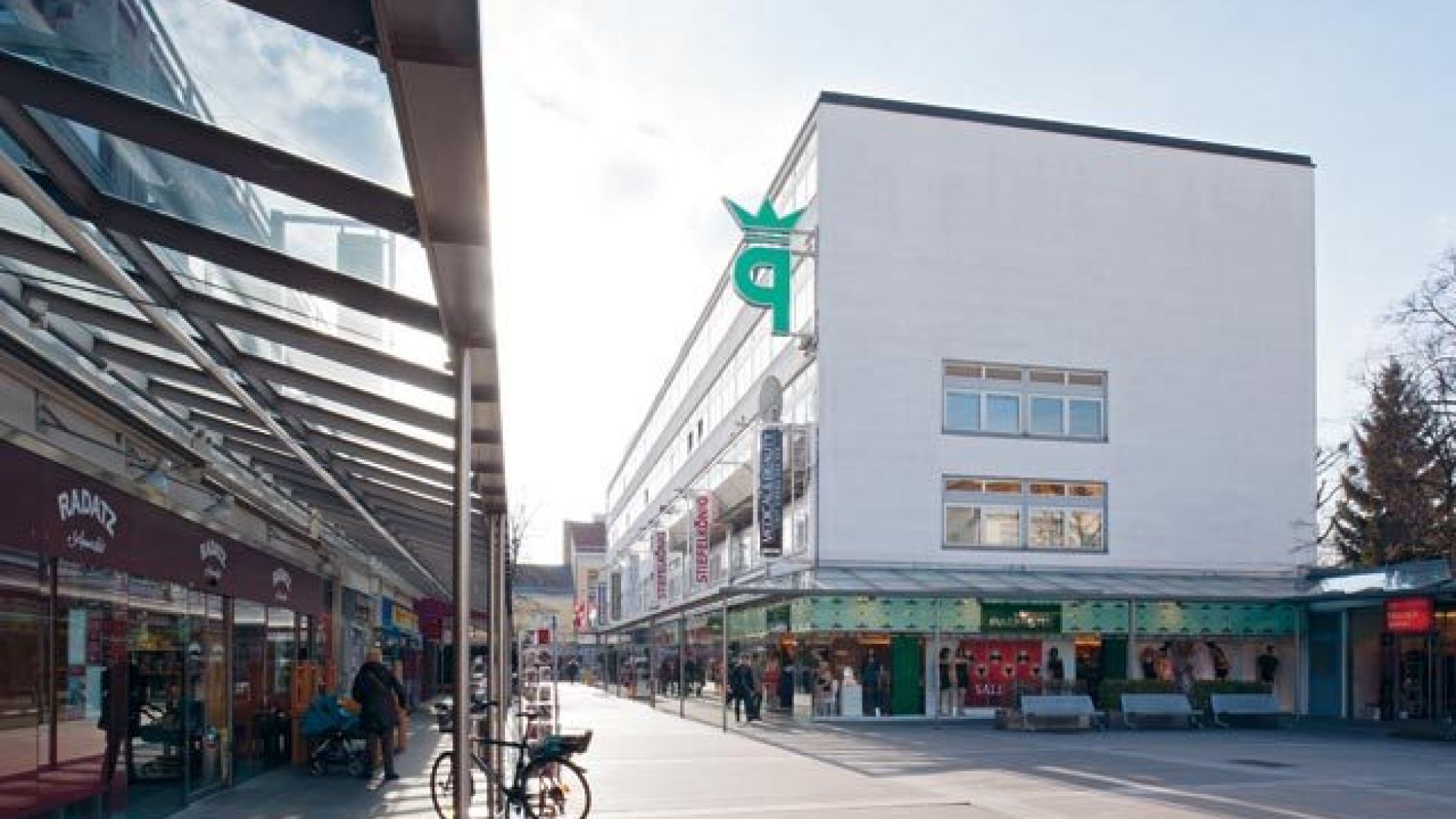 copyright: ©Pez Hejduk Blick unter einem Glasvordach entlang eines länglichen Gebäudes, rechts parallel dazu ein weiteres Gebäude