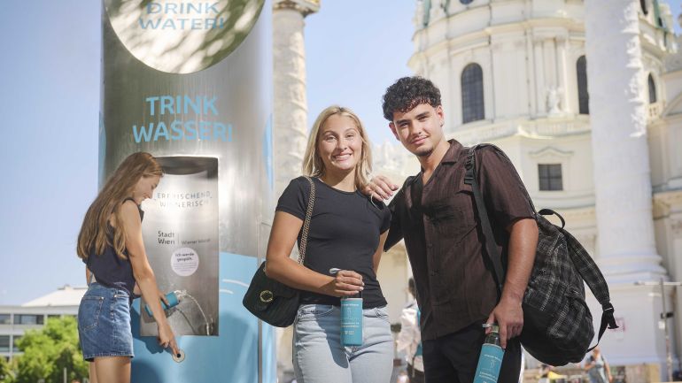 copyright: Wiener Wasser/Zinner Zwei junge Leute stehen mit Wasserflaschen vor dem Trinkbrunnen vor der Karlkirche. Eine andere junge Frau füllt ihre Flasche mit Wasser auf.