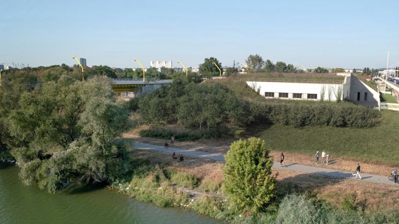 copyright: Stadt Wien/convex/Pulse/Illichmann/Janusch Zwischen 2 Brücken auf der Donauinsel steht ein niedriges weißes Gebäude mit begrüntem Dach. Davor wachsen Sträucher. Am Gehweg am Ufer spazieren Menschen.