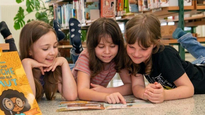 copyright: Stadt Wien - Büchereien/Natalie Stephan Drei Mädchen liegen in einer Bücherei auf dem Boden und lesen ein Heft.