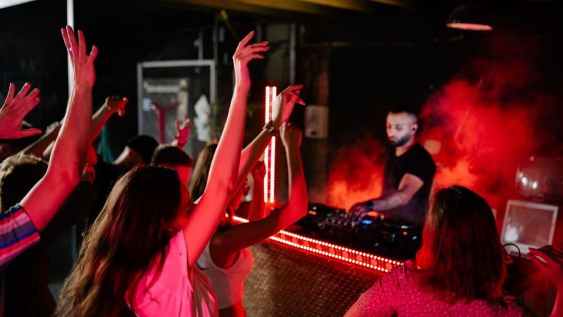 copyright: Pexels/Yankrukov Tanzende Party-Gäste vor einem DJ am DJ-Pult mit rotem Rauch im Hintergrund.