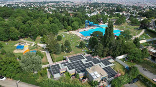 Blick von oben auf das Schafbergbad: Gebude mit Photovoltaik-Anlagen auf dem Dach, Sandspielplatz, Liegewiese, Schwimmbecken im Freien mit groer, blauer Rutsche