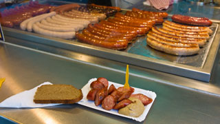 Wrstel mit Senf und Brot stehen auf der Theke, im Hintergrund braten weitere Wrstel