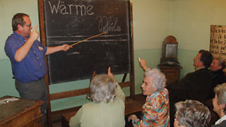Group of elderly people in an antiquated schoolroom: a man pointing at the black board with a stick.