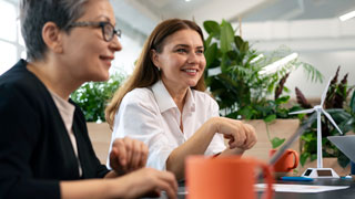 2 Frauen sitzen an einem Schreibtisch in einer Besprechung; vor ihnen am Tisch ein Laptop