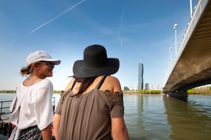 Zwei Jugendliche sitzen an der Donau mit Blick auf DC-Tower neben der Reichsbrücke