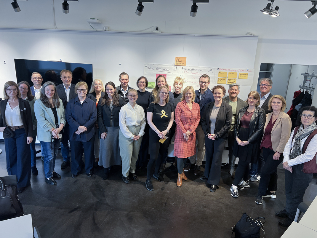 Gruppenfoto von etwa 20 Personen in einem modernen Besprechungsraum, die an einem Workshop teilnehmen. Teilnehmer tragen überwiegend formelle Kleidung, einige lächeln, und die Atmosphäre wirkt freundlich und kooperativ.