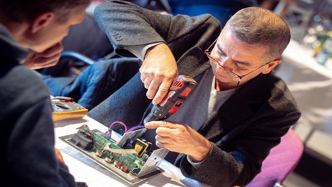 The photo shows a person working on an open electronic circuit board with a cordless screwdriver.