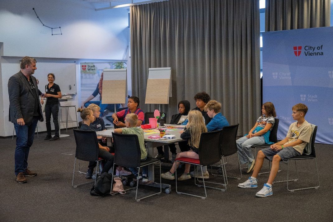 Fotografie einer Gruppensituation mit Kindern und Günther Siebenhaar in einem Seminarraum, erkennbar an Bannern der Stadt Wien und City of Vienna. Kinder sitzen um einen Tisch mit Bastelmaterialien, während Günther Siebenhaar spricht und eine Frau im Hintergrund lächelt.