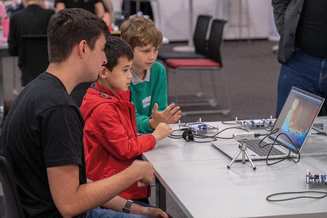 Fotografie zeigt drei Personen, darunter zwei Kinder, die an einem Tisch mit elektronischen Bauteilen und einem Laptop sitzen, bei einer technischen Lern- bzw. Programmieraktivität. Laptop-Bildschirm zeigt farbige Grafiken.