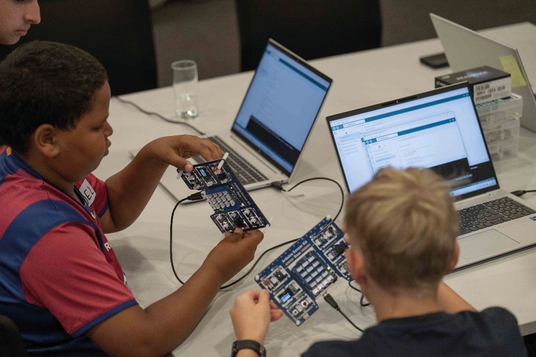 Fotografie zeigt zwei Kinder, die an einem Tisch mit Laptops und elektronischen Schaltkreisen arbeiten. Fokus liegt auf praktischer Anwendung von Elektronik, erkennbar an den Platinen und geöffneten Programmierumgebungen auf Bildschirmen.