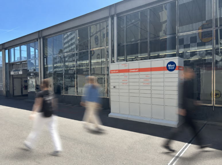 Auf einem Foto ist eine Next Box abgelichtet worden. Diese Box steht vor einer U-Bahnstation. Menschen werden schemenhaft dargestellt, wie sie vorbei gehen.
