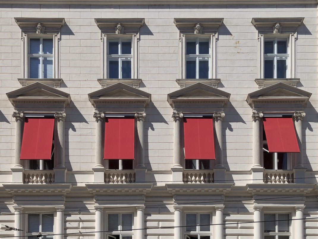 Fallarmmarkisen an einem dekorierten Gründerzeithaus in einer Schutzzone. Der eckige Vorbaukasten findet in der Fensterlaibung der Kastenfenster Platz, die Konstruktion wird in Fensterfarbe ausgeführt. Der Textilbehang ist farbig gestaltet. Die Sonnenschutzvorrichtungen an den einzelnen Fenstern des Hauses sind einheitlich nach diesem Prinzip gestaltet.