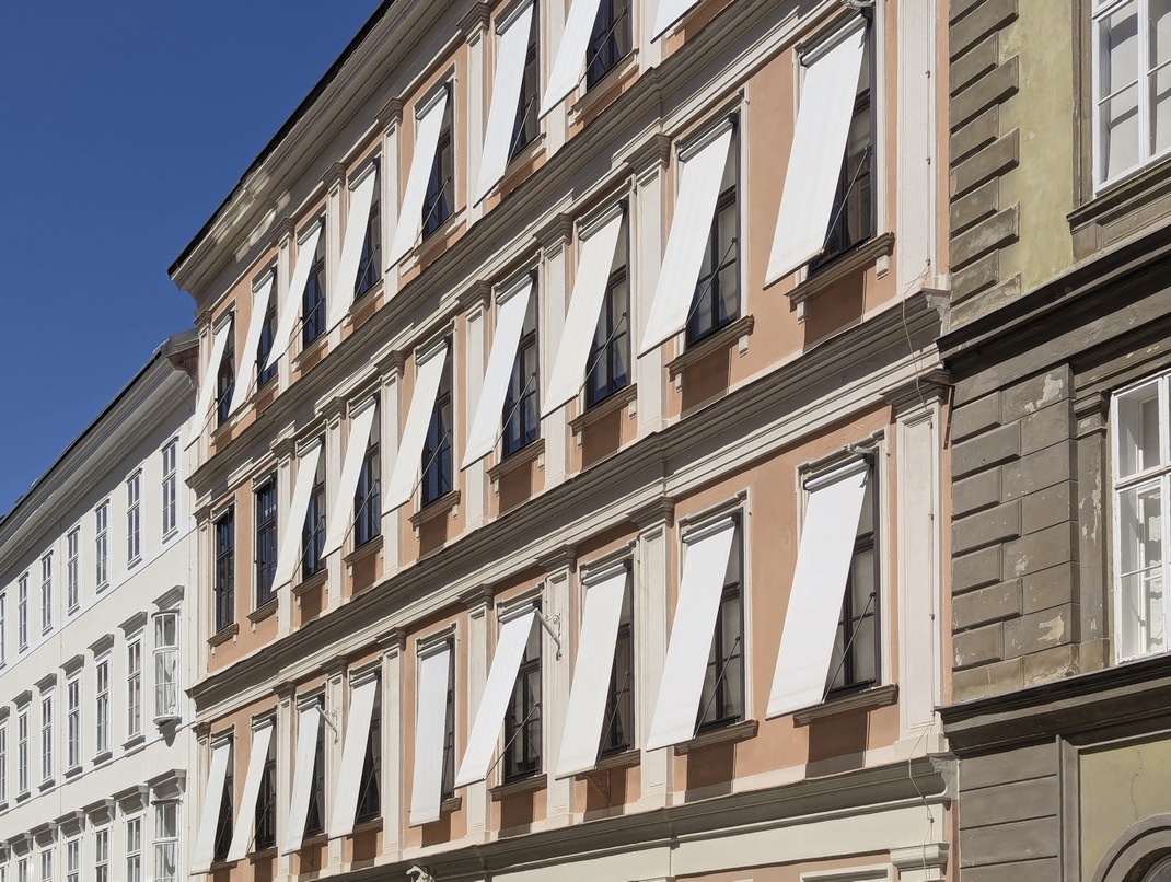 Fallarmmarkisen an einem dekorierten Gründerzeithaus in einer Schutzzone. Die Markisenrolle wird ohne Vorbaukasten vor dem Kastenfenster angebracht, die Konstruktion wird in Fensterfarbe ausgeführt. Der Textilbehang ist weiß gestaltet. Der Sonnenschutz ist an der Fassade einheitlich gestaltet.