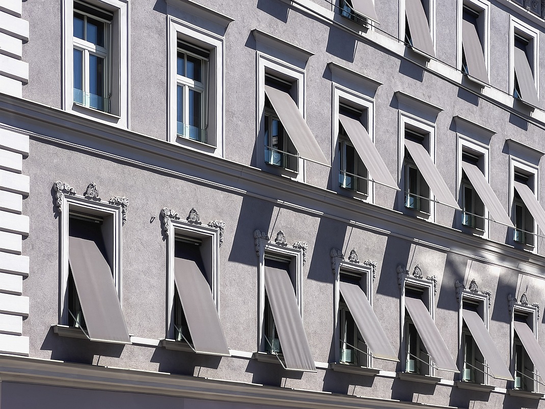 Bewilligungsfreie Fallarmmarkisen an einem dekorierten Gründerzeithaus außerhalb von Schutzzonen. Der eckige Vorbaukasten findet in der Fensterlaibung Platz, die Konstruktion wird in Fensterfarbe ausgeführt. Der Textilbehang ist in Fassadenfarbe gestaltet. Der Sonnenschutz ist an der Fassade einheitlich gestaltet.