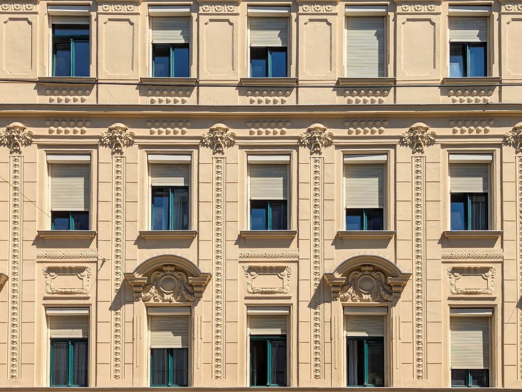 Bewilligungsfreie Rollläden an einem dekorierten Gründerzeithaus außerhalb von Schutzzonen. Der eckige Vorbaukasten findet in der Fensterlaibung Platz, die Konstruktion sowie der Rollladen werden in Fassadenfarbe gestaltet. Der Sonnenschutz ist an der Fassade einheitlich gestaltet.