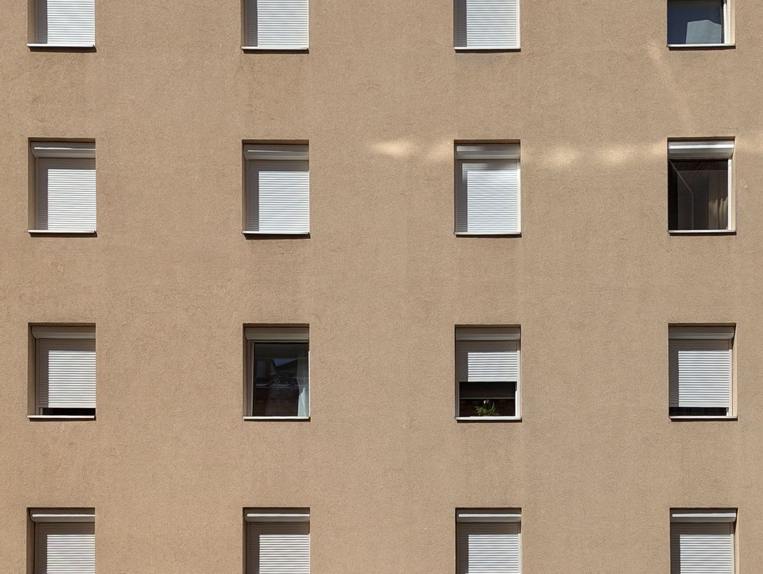 Bewilligungsfreie Rollläden an einem Wohngebäude der Nachkriegszeit mit glatter Fassade außerhalb von Schutzzonen. Der eckige Vorbaukasten findet in der Fensterlaibung Platz, die Konstruktion sowie der Rollladen werden in Fensterfarbe gestaltet. Der Sonnenschutz ist an der Fassade einheitlich gestaltet.