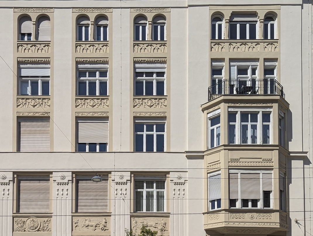 Rollläden an einem dekorierten Gründerzeithaus in einer Schutzzone. Der eckige Vorbaukasten findet in der Fensterlaibung der Kastenfenster Platz, die Konstruktion sowie der Rollladen werden in Fensterfarbe gestaltet. Der Vorbaukasten besitzt Ornamentik. Der Sonnenschutz ist an der Fassade einheitlich gestaltet.