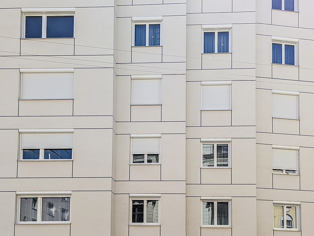 Bewilligungsfreie Rollläden an einem Wohngebäude der Nachkriegszeit mit glatter Fassade. Der eckige Vorbaukasten findet in der Fensterlaibung Platz, die Konstruktion sowie der Rollladen werden in Fensterfarbe gestaltet. Der Sonnenschutz ist an der Fassade einheitlich gestaltet.