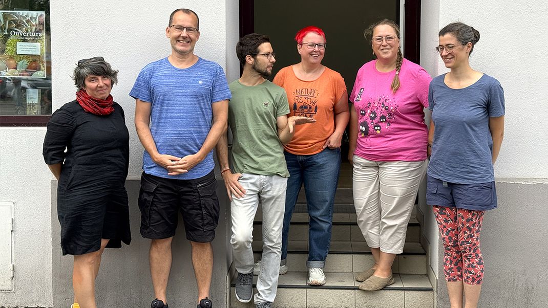 Six young and middle-aged people, dressed casually, are standing in a row, looking relaxed and cheerful, in front of a shopfront bearing the sign ‘Solidar:Raum’