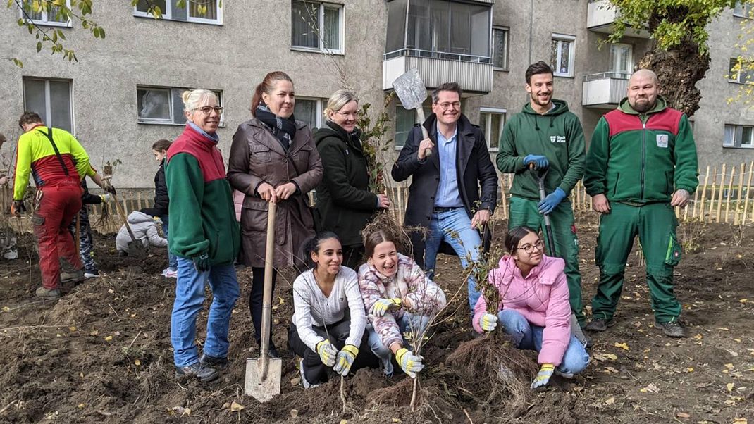 On a ploughed-up patch of land in front of a block of council flats, a group of adults is standing in the second row, spades in hand; in front of them, three children are crouching, holding plants.