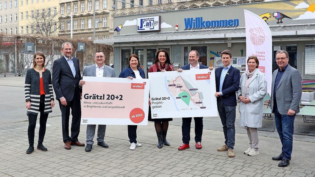 Nine people are standing in a row, smiling at the camera. They are holding two large signs referring to the Grätzl 20+2 district and showing a map section indicating its geographical location. Behind them is a low-rise building bearing the inscription ‘Welcome’, as well as buildings from the Gründerzeit period.