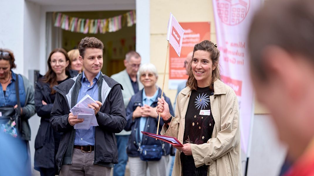 A group of people. In the centre-right, a young woman (Helene Scheller) holding a small flag is smiling (Helene Scheller), and to her left, a young man (Stephan Hartmann) is looking at her. In the background, other people are watching the scene unfold. In the background, the entrance to a ground-floor pub.