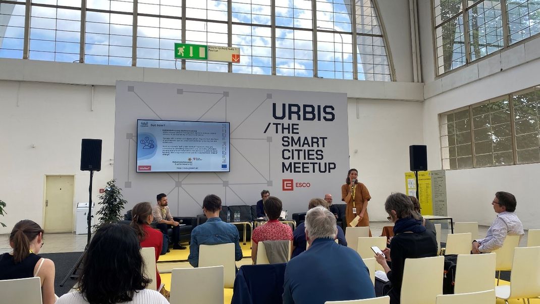 A photo from the conference showing Helene Scheller on stage in front of the audience, beneath the conference poster ‘URBIS / The smart Cities Meetup’.