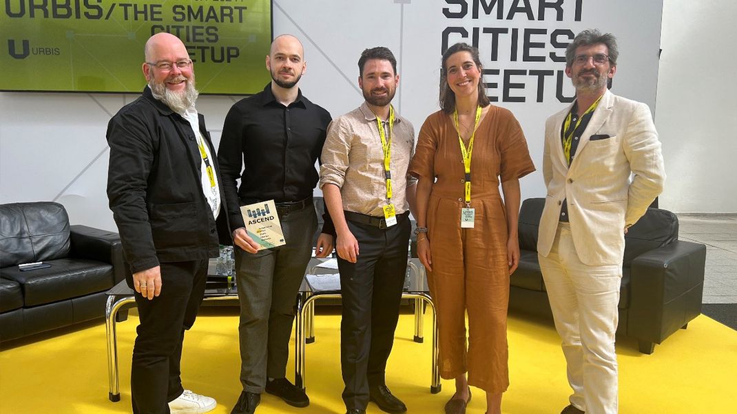 Four men and a woman (Helene Scheller), with the woman in the centre looking at the camera; in the background, the logo of the URBIS conference. Far left Bernhard Klassen (Munich), far right Maxime Valentin (SPL Lyon Confluence)