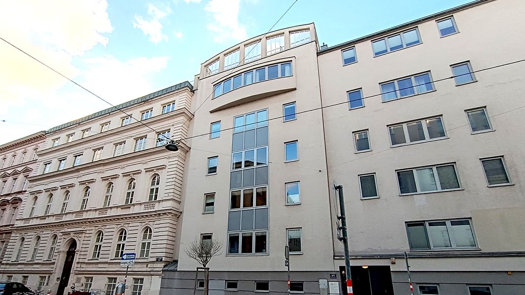 A view of the façades of two residential buildings, one with four storeys and the other with five; on the left, a Gründerzeit-style building with a structured façade and a converted attic; on the right, a more modern building with a smooth façade and a striking bay window. Both façades are white-beige.
