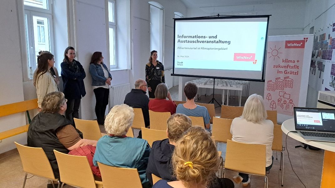 An information session with a large projection screen in the background, with people seated in front of it. Some are standing at the edge of the room.