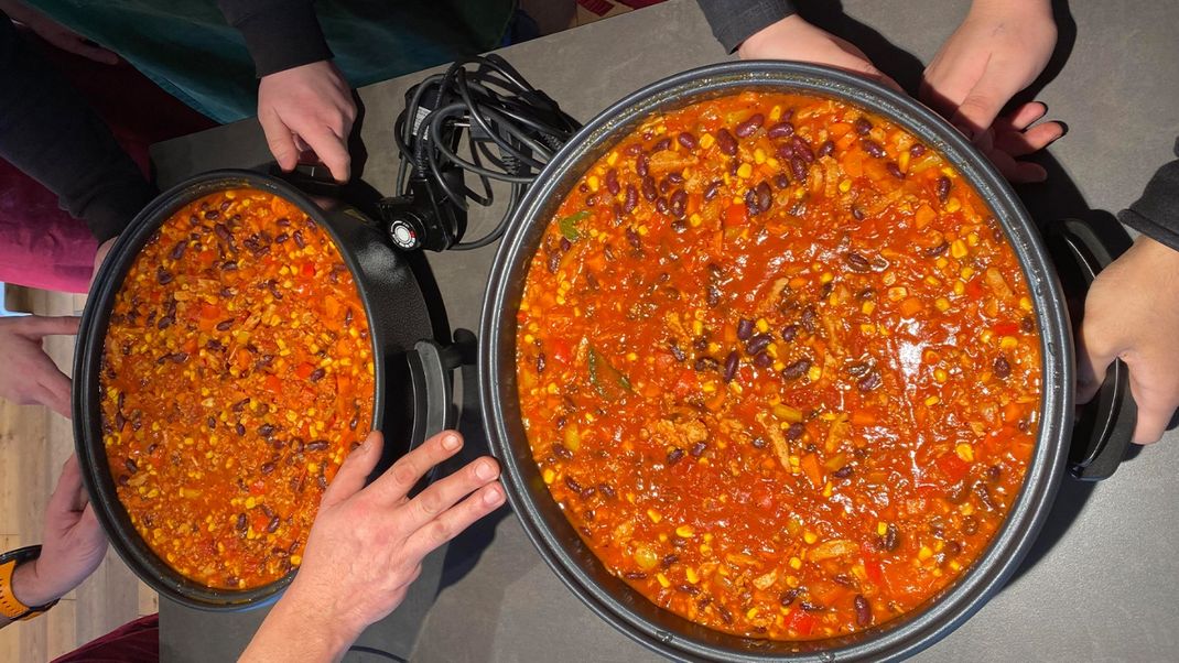The picture shows two full roasting tins containing a red-orange bean dish. Several hands are holding them.