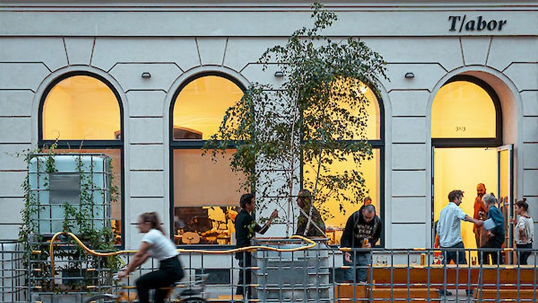 A bustle of activity in a brightly lit ground-floor premises with four tall arched windows. In front of it stands a tree and various stands, suggesting there are benches in front of them.