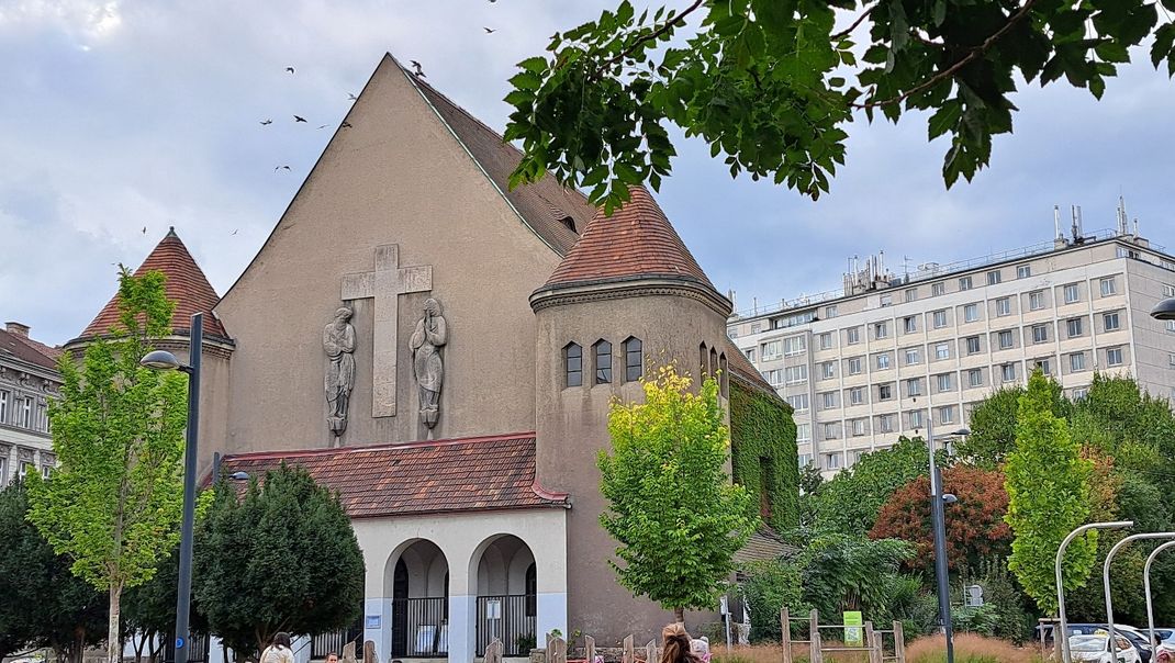 A photograph of the main façade of the Protestant church with a gabled roof; in the pointed gable is a cross flanked by two figures. To the left and right are small towers, in front of which is the porch with arcades. The church is surrounded by trees, with a modern residential high-rise in the background.