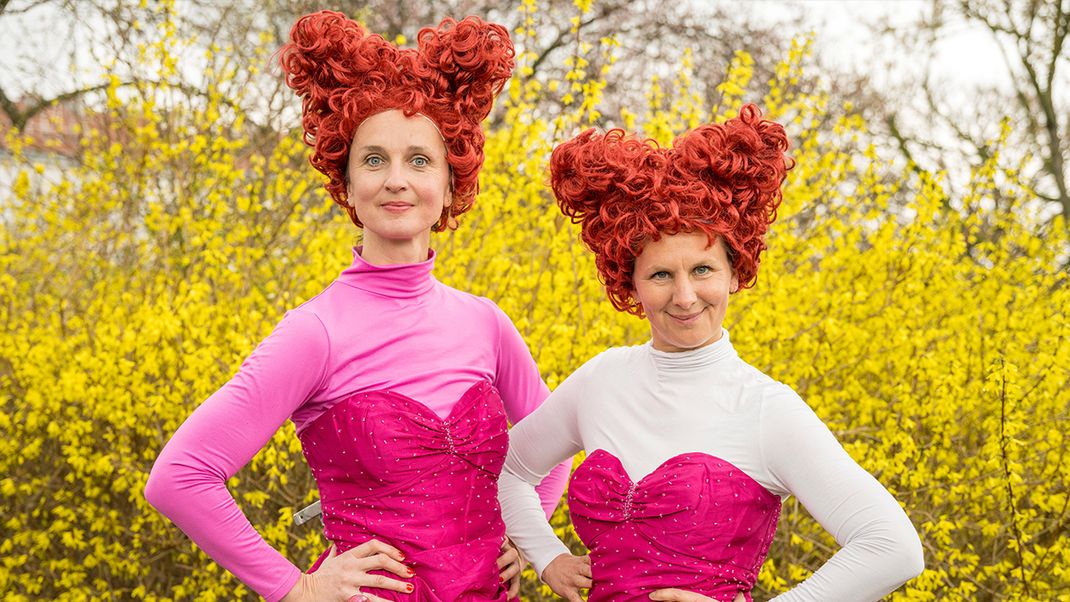 Two fairy-tale characters dressed in crimson, with orange hair standing on end, against a yellow forsythia background.