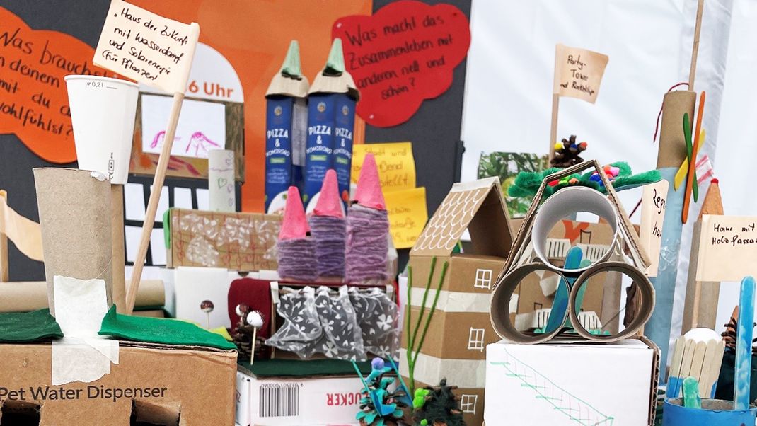 Bunte Kinderbastelei mit Kartonschachteln, Garnen und Papier, die eine Stadt mit in der Mitte einer Straße darstellt.