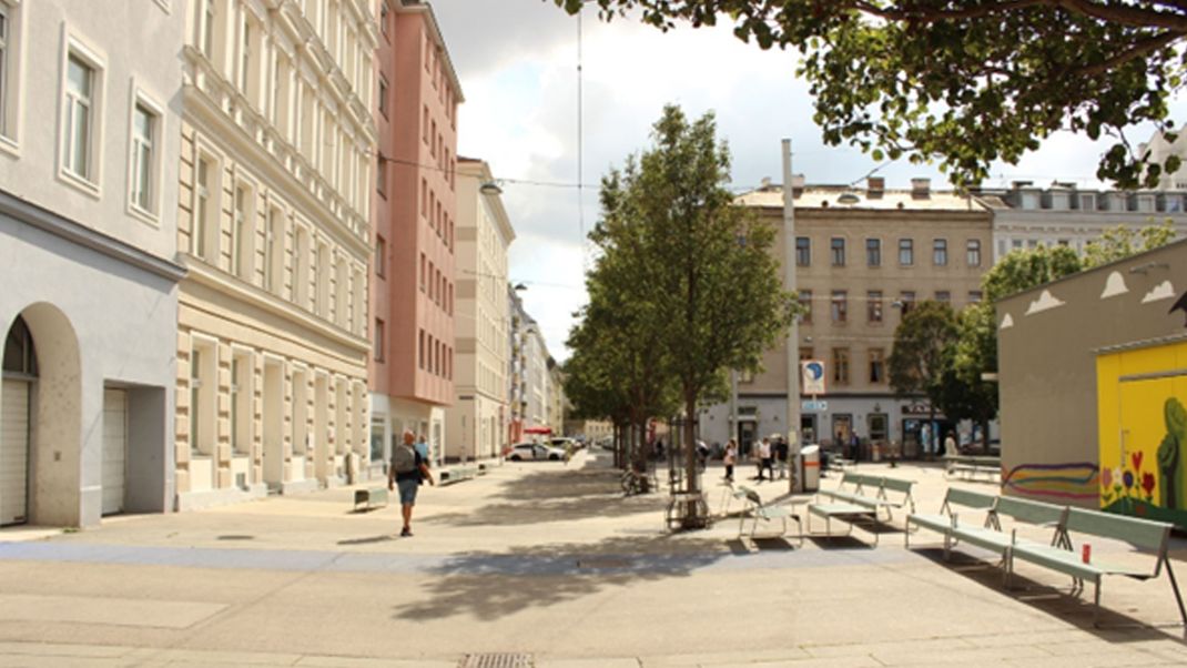 Urbaner Raum mit Altbauten links, einer hell gepflasterten Freifläche in der Mitte sowie mit Bäumen und Bänken in der Mitte und rechts.